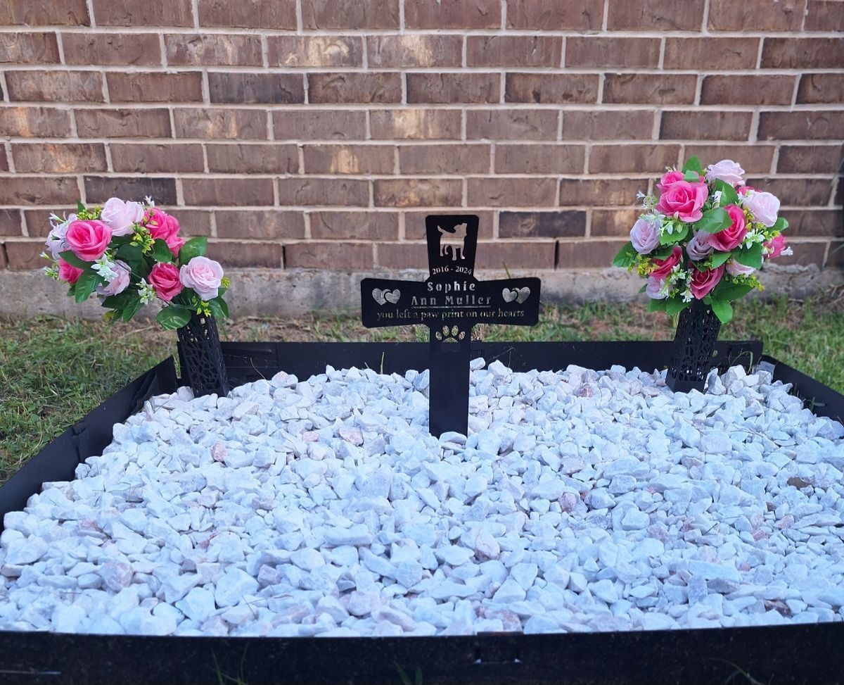 Pet memorial with engraved plaque and flower arrangements on white stones against a brick wall.